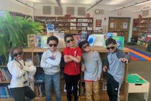 A group of elementary-aged boys and girls wearing sunglasses are posing in front of books in a school library