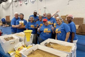 A group of men and women are wearing hair nets and bright blue shirts while smiling at the camera. They are in a gym, standing behind an assembly line that includes large boxes of dry macaroni and soy.