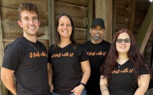 Two men and two women are standing in front of the wooden slats of a covered bridge. They are wearing black t-shirts that say It starts with U in orange