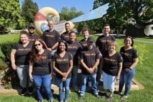 A group of men and women of varying ages are standing in a group outside. It is a sunny day and there is a distlefink statue behind them. They are all wearing black t-shirts and jeans with the words It starts with U written on the front of their shirts